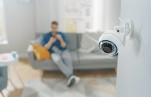 Close-up-shot-of-a-home-security-camera-on-a-wall-in-an-apartment.-A-man-is-sitting-on-a-sofa-in-the-background.-Credit_gorodenkoff_istock.com