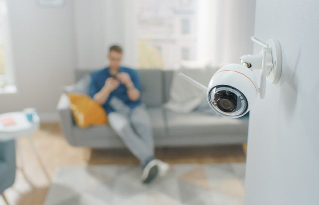 Close up shot of a home security camera on a wall in an apartment. A man is sitting on a sofa in the background. Credit: gorodenkoff/ iStock.com